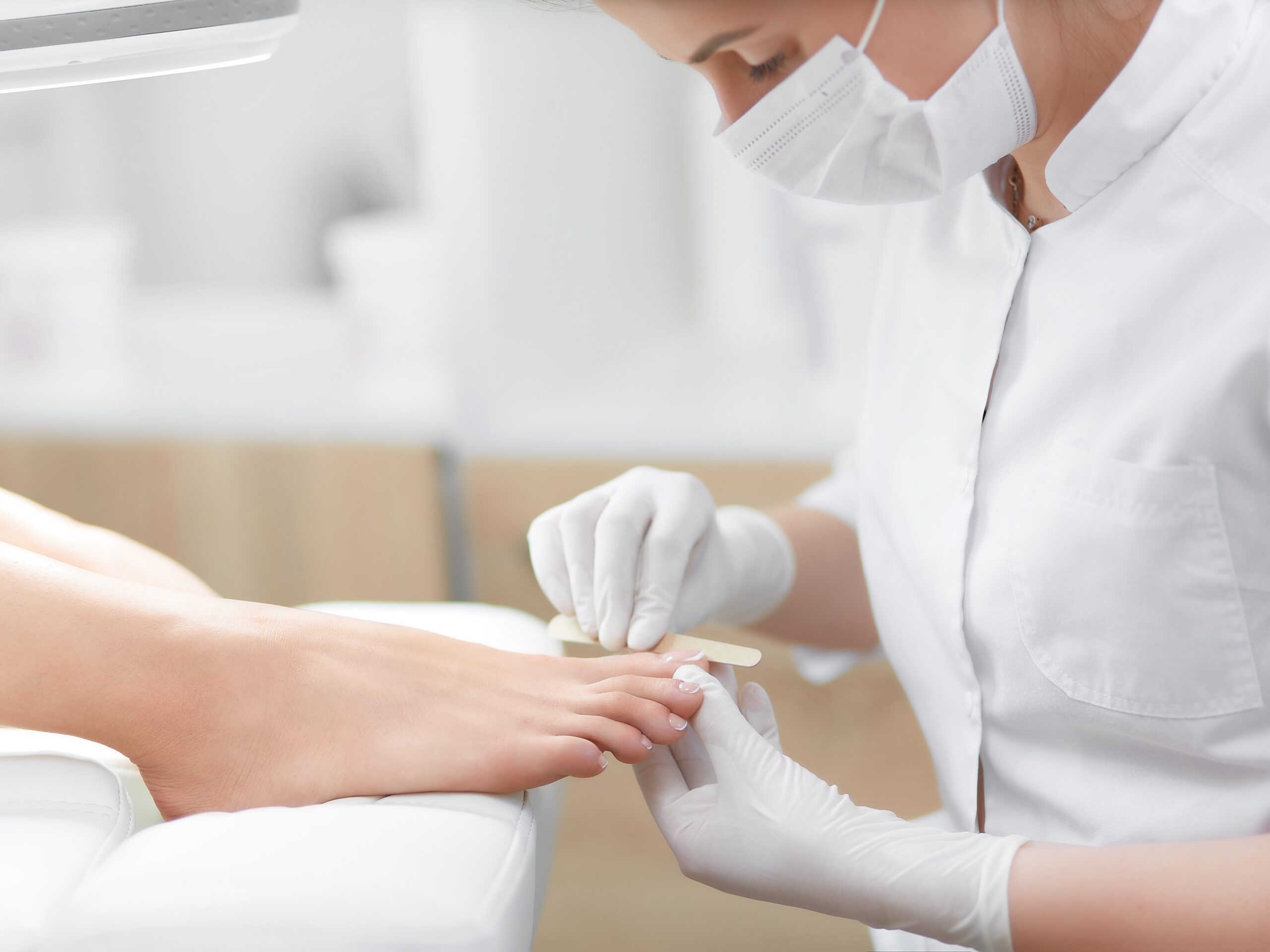 Nail artist in beauty salon making french pedicure for client`s feet. Professional in mask on face polish nails by tool. Concept of nails, feet health and podiatrist proffeshion.